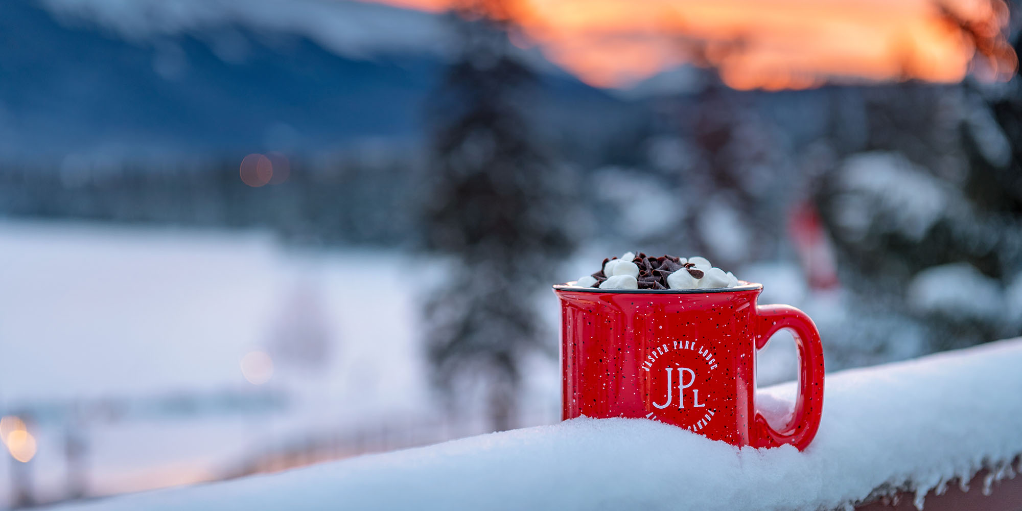 A red JPL mug of hot chocolate sits on a snowy ledge with the sun setting behind mountains in the background.
